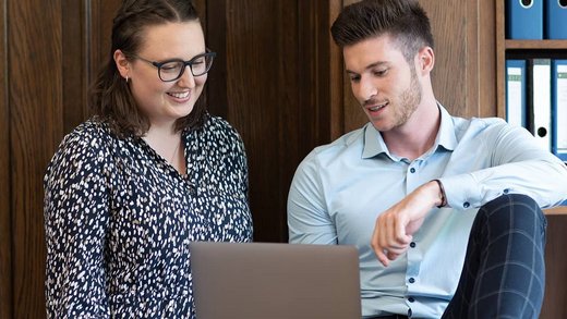 Eine Kollegin und ein Kollege sitzen lässig auf einer Mauer und schauen in einen Laptop