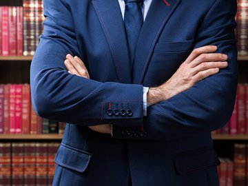 Compliance-Management: Ausschnitt Mann im blauen Anzug, vor dem Oberkörper verschränkte Arme, Bücherregal im Hintergrund