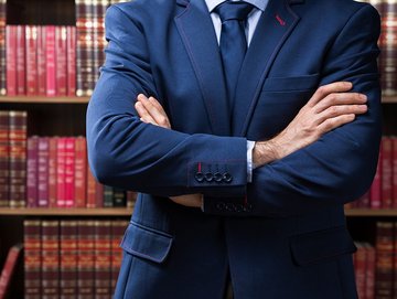 Compliance-Management: Ausschnitt Mann im blauen Anzug, vor dem Oberkörper verschränkte Arme, Bücherregal im Hintergrund