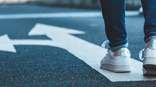 Global Mobility Services – internationaler Mitarbeitereinsatz: Beine einer Person, die auf einer Straße läuft auf der ein Pfeil geradeaus und nach rechts zeigt