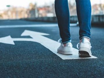 Global Mobility Services – internationaler Mitarbeitereinsatz: Beine einer Person, die auf einer Straße läuft auf der ein Pfeil geradeaus und nach rechts zeigt